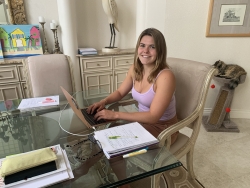 Jennifer Secrest sitting in front of a laptop at a dining room table in her home. There are notebooks on the table, and a cat sitting on a cat tree in the background.